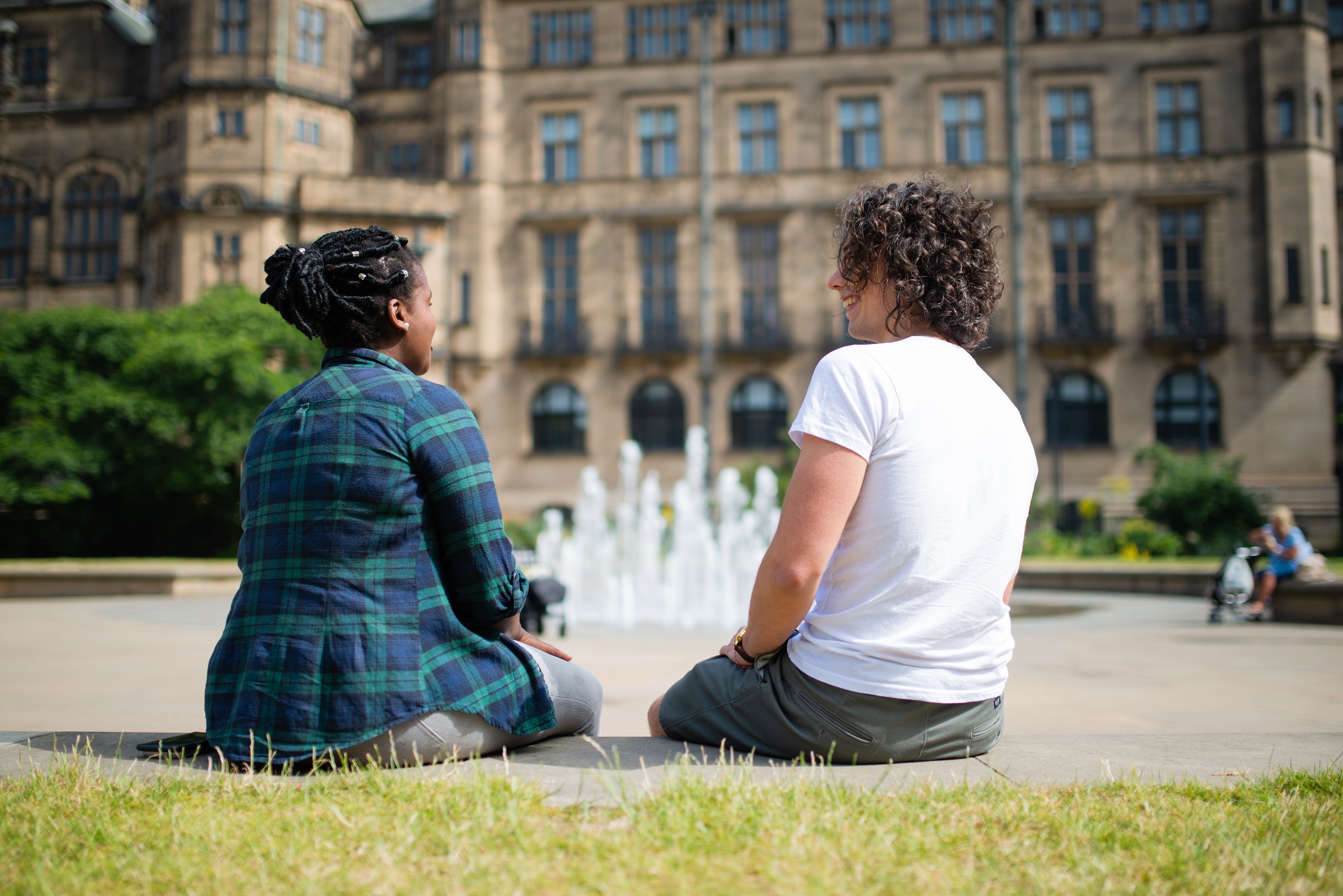 Two students sitting outside a prestigious looking building and a fountain. The two students have their back to the camera. 