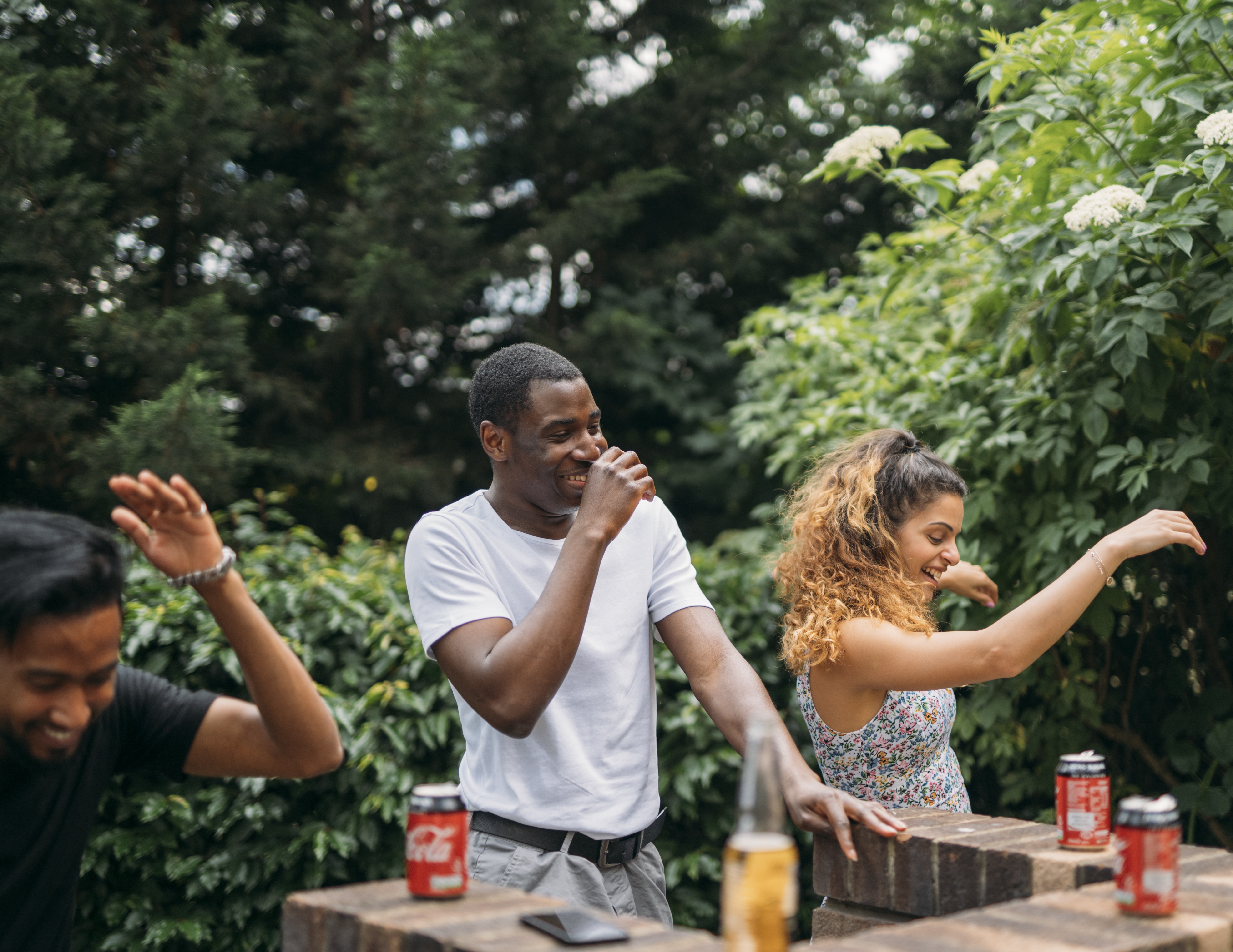 Celebrate friendships - three friends are laughing and cheering out in a sunny garden, with beers and coke zero in front of them. 